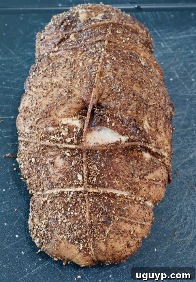 A fully prepared turkey breast, tightly secured with butcher's twine, lies on a sleek black cutting board. Its surface is completely enveloped in a thick, dark, and textured spice rub, showcasing the rich seasoning before cooking.