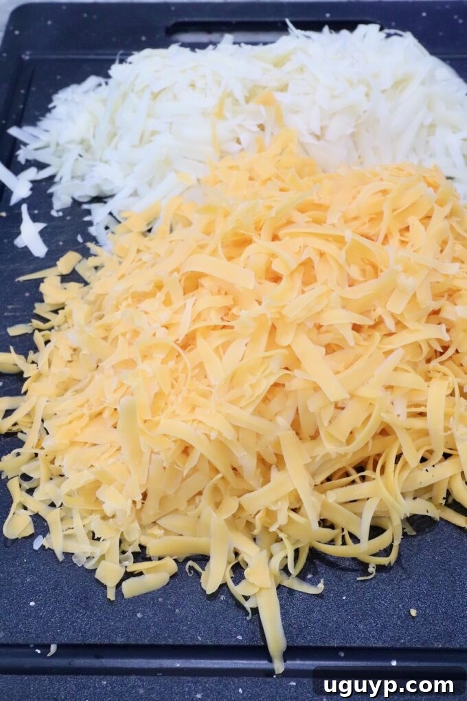 A pile of finely grated swiss cheese and a separate pile of cheddar cheese are on a black cutting board