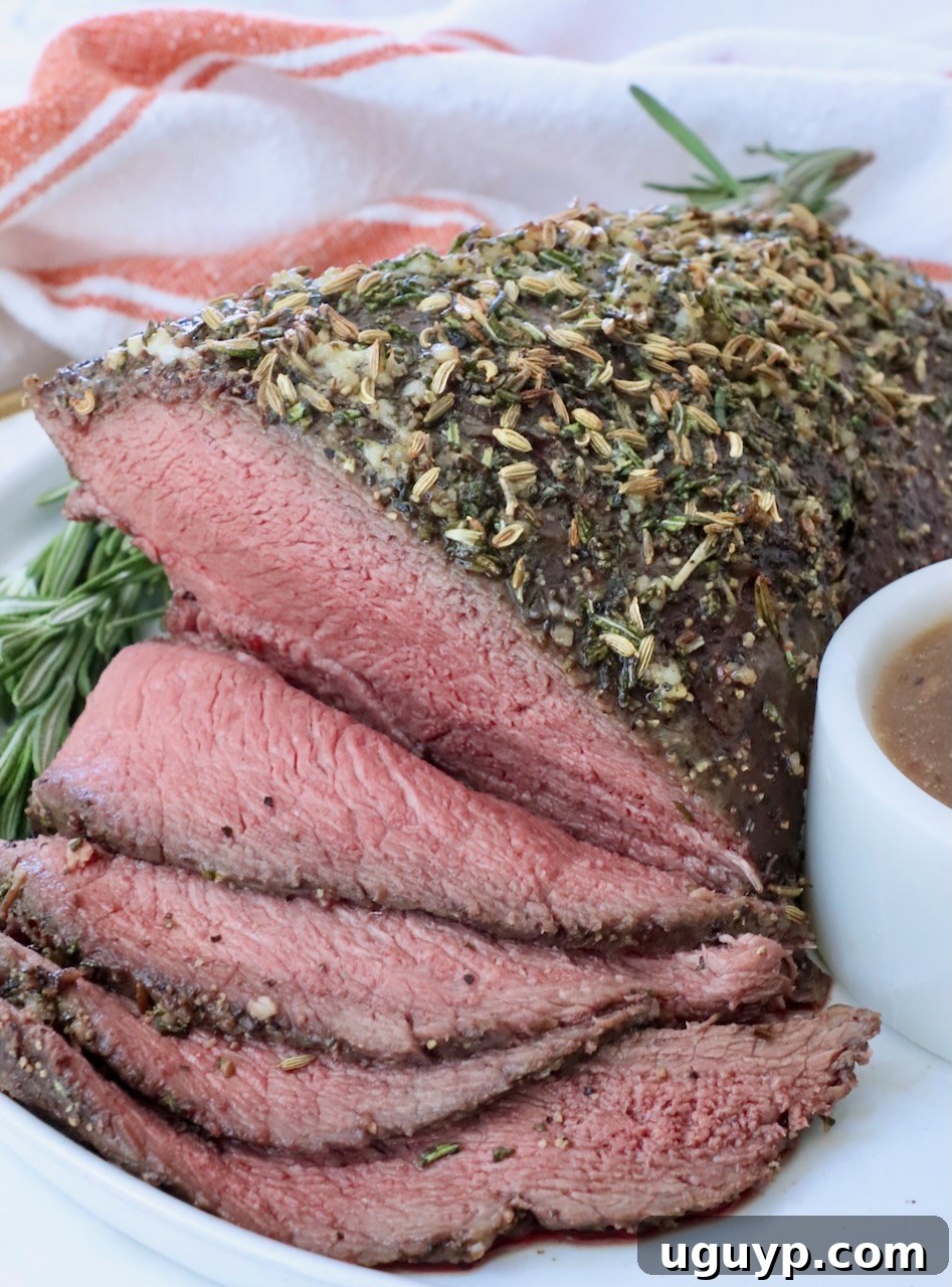 Roast beef topped with herbs and spices, sliced to reveal a juicy pink interior. Garnished with rosemary and served on a white plate with gravy.