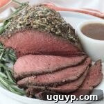 Roast beef topped with herbs and spices, sliced to reveal a juicy pink interior. Garnished with rosemary and served on a white plate with gravy.