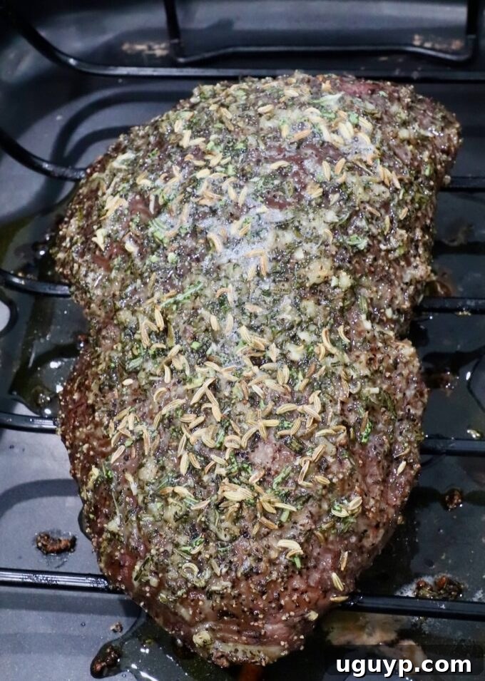 A seasoned roast beef resting on a wire rack, topped with herbs and spices, in a dark roasting pan