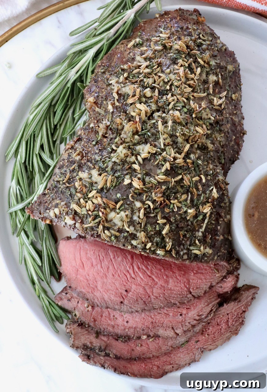 Roasted beef topped with herbs, sliced on a white plate with a sprig of rosemary. A small bowl of gravy sits on the side. 