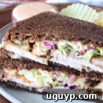 Close-up of a sandwich with dark rye bread filled with sliced turkey, creamy slaw, and sauce. Served on a plate with wavy potato chips and a dipping sauce.