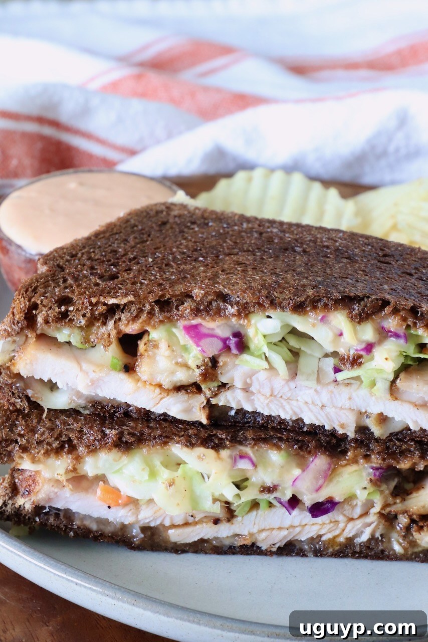 Close-up of a Rachel sandwich with dark rye bread filled with sliced turkey, creamy slaw, and sauce. Served on a plate with wavy potato chips and a dipping sauce.