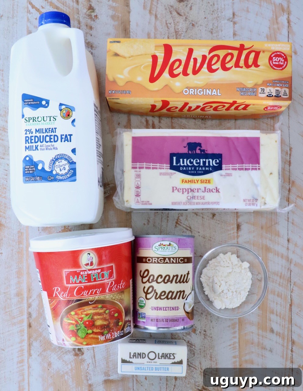 A flat lay of cooking ingredients on a wooden surface. Items include milk, Velveeta cheese, pepper jack cheese, red curry paste, coconut cream, flour, and a stick of butter.