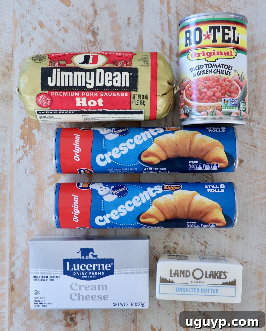 Assorted grocery items on a wooden surface: Jimmy Dean pork sausage, Rotel tomatoes, two Pillsbury crescent roll cans, Lucerne cream cheese, Land O'Lakes butter.