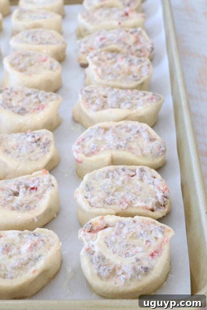 Rows of uncooked pastry pinwheels filled with a creamy mixture of sausage and tomatoes are neatly arranged on a parchment-lined baking sheet.