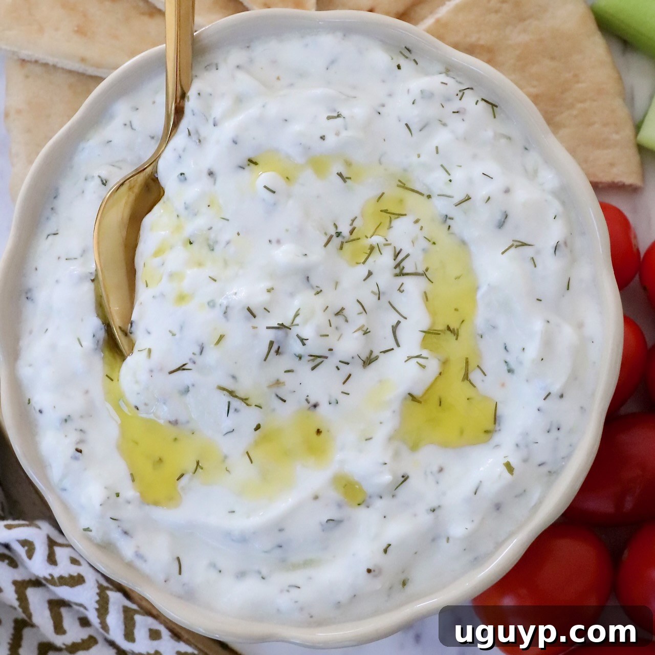 tzatziki sauce in a bowl with a spoon with olive oil drizzled on top and slices of pita bread and cherry tomatoes on the side
