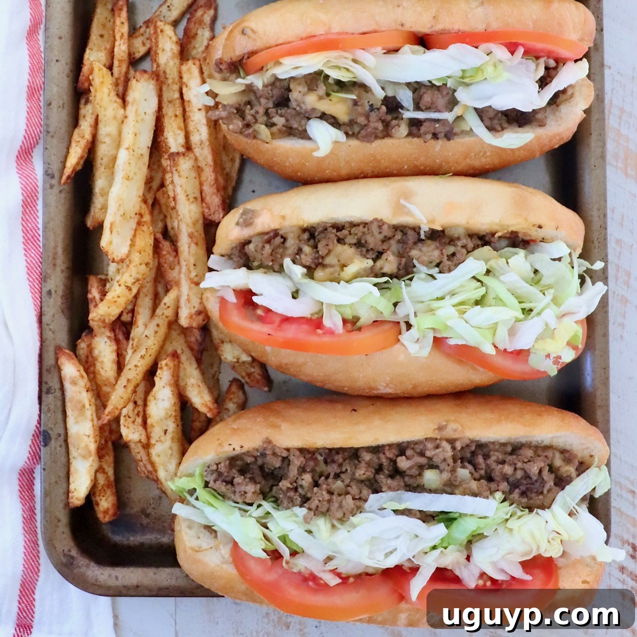 three chopped cheese sandwiches with sliced tomatoes and shredded lettuce lined up on a baking sheet with seasoned, cooked french fries on the side