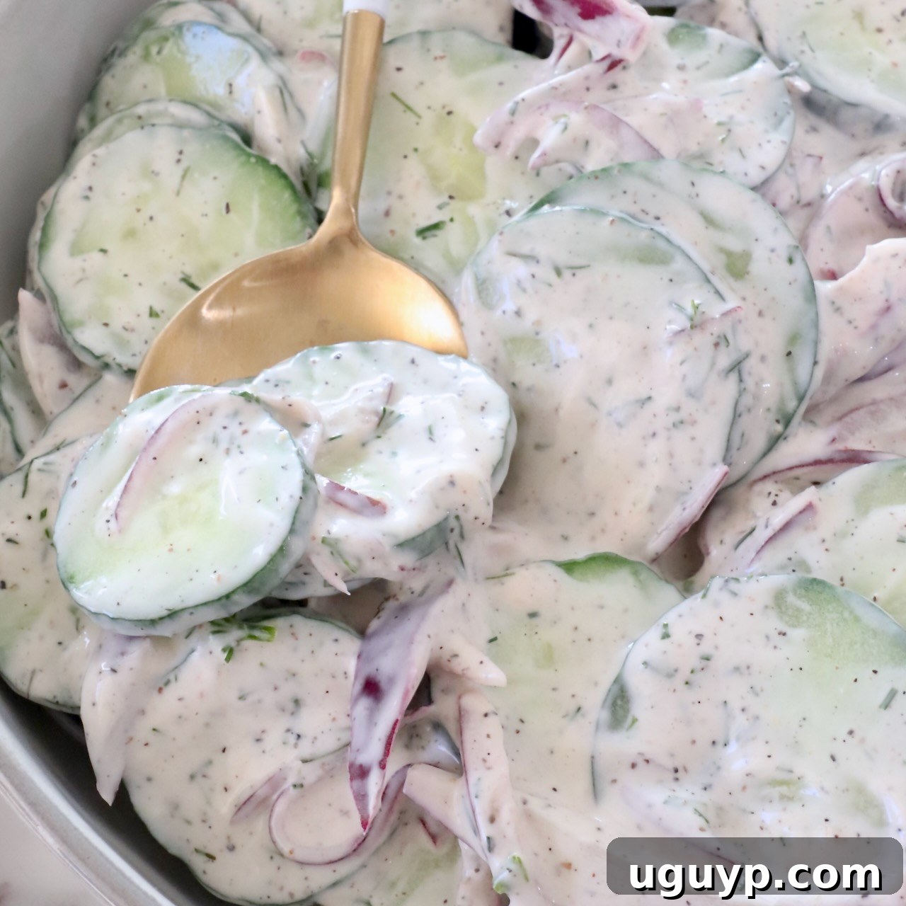 sliced cucumbers and red onions tossed in a creamy dressing in a bowl with a gold spoon