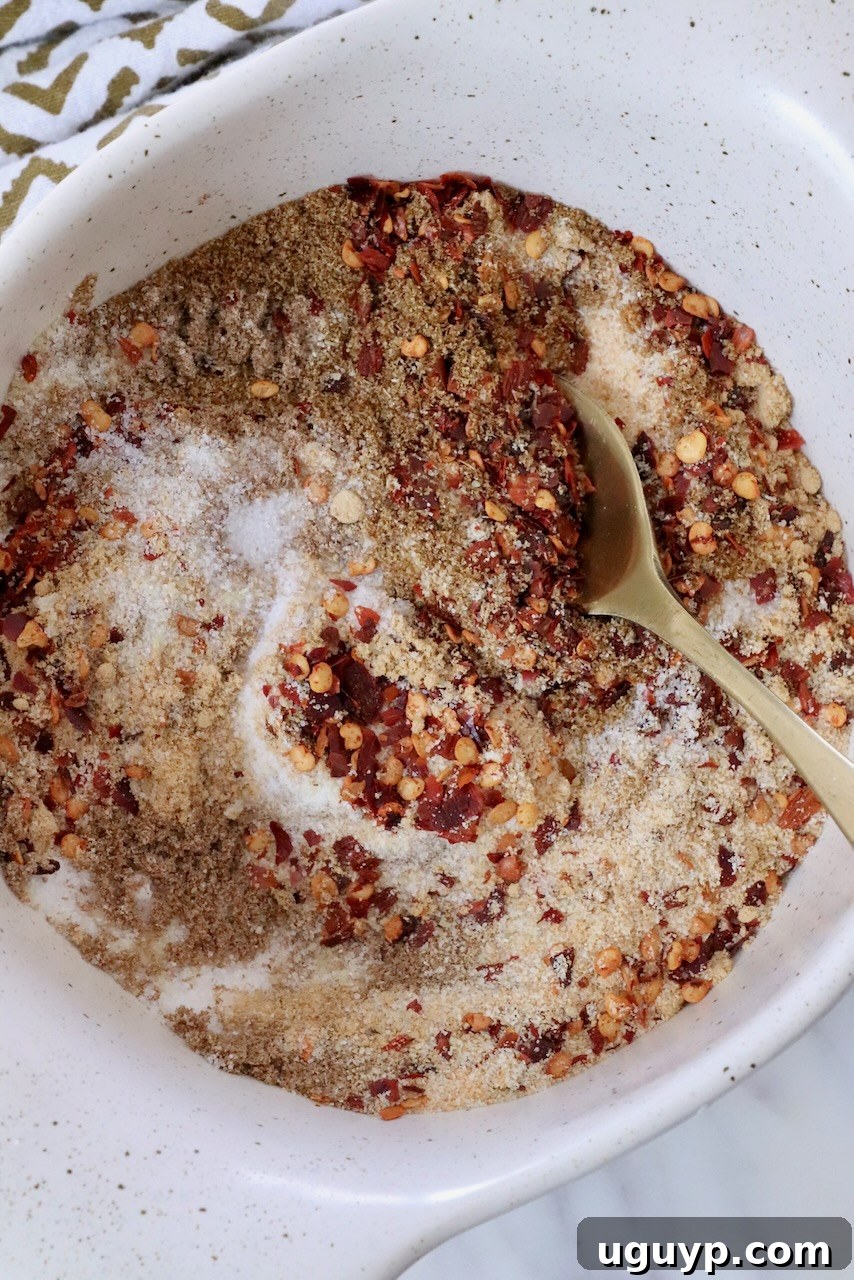 A white bowl contains a mix of red chili flakes, brown spices, and white salt, stirred with a golden spoon. A patterned cloth is partially visible.