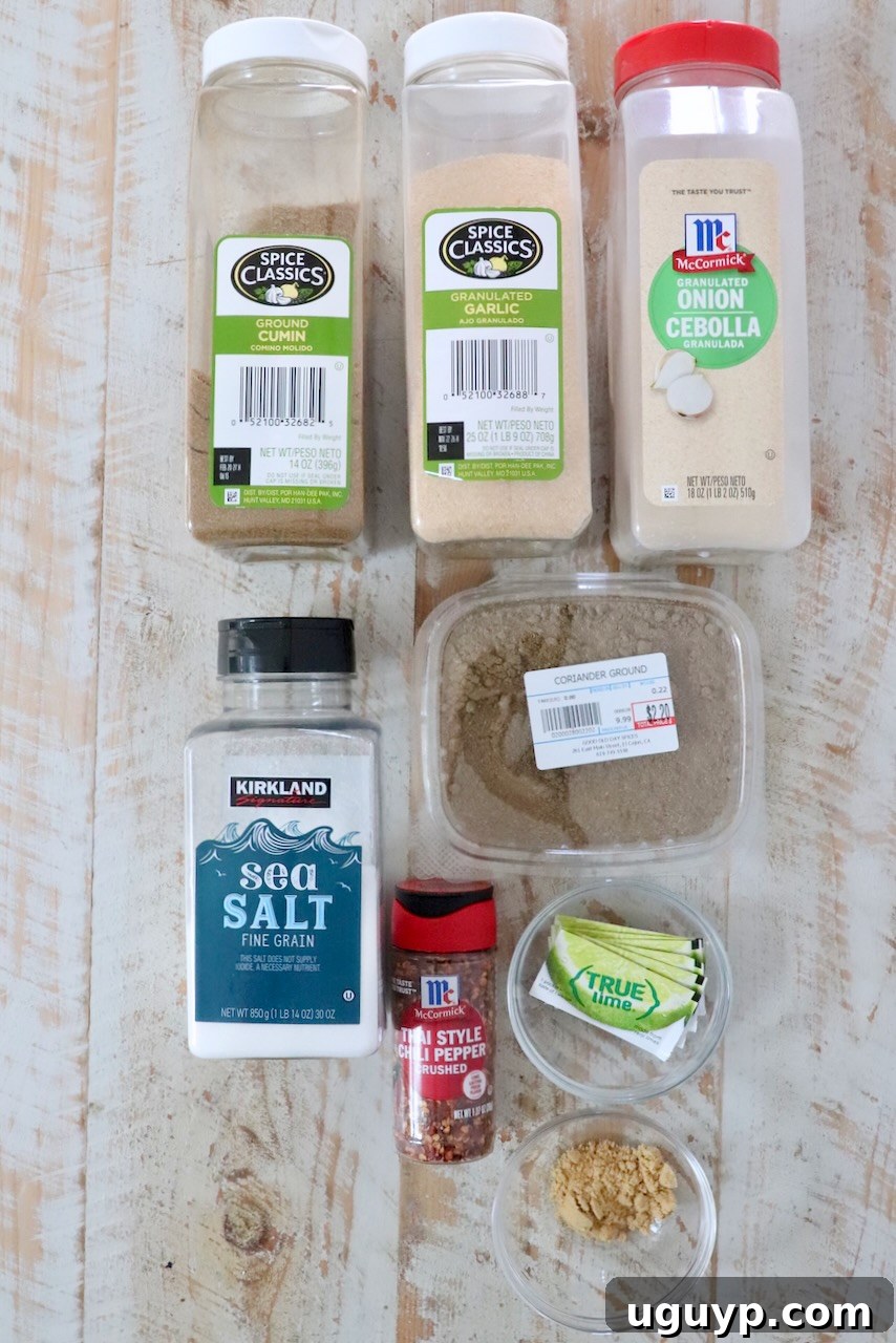 Various spice containers arranged on a wooden surface: cumin, garlic, onion powder, sea salt, ground coriander, chili flakes, lime packets, ground ginger.