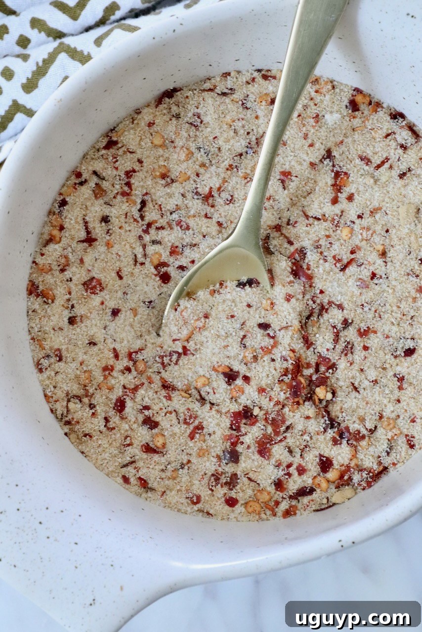 A white bowl filled with a dry mixture of salt, spices and red pepper flakes, featuring a metal spoon. A patterned cloth is partially visible beside the bowl.