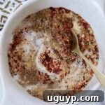 A white bowl contains a mix of red chili flakes, brown spices, and white salt, stirred with a golden spoon. A patterned cloth is partially visible.