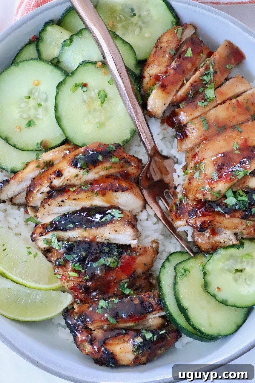 Grilled, sliced Thai chicken with a glossy glaze on a bed of fluffy white rice, garnished with fresh herbs. It is served on a pristine white plate alongside crisp cucumber slices and bright lime wedges, with a fork ready for enjoyment.