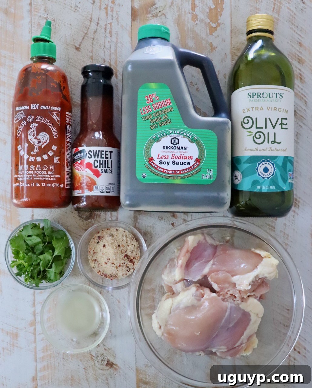 Fresh ingredients for Grilled Thai Chicken arranged on a rustic wooden surface. From left to right: a bottle of Sriracha, a jar of sweet chili sauce, a bottle of soy sauce, olive oil, fresh cilantro sprigs, a bowl of Thai seasoning, sliced lime wedges, and raw boneless, skinless chicken thighs.