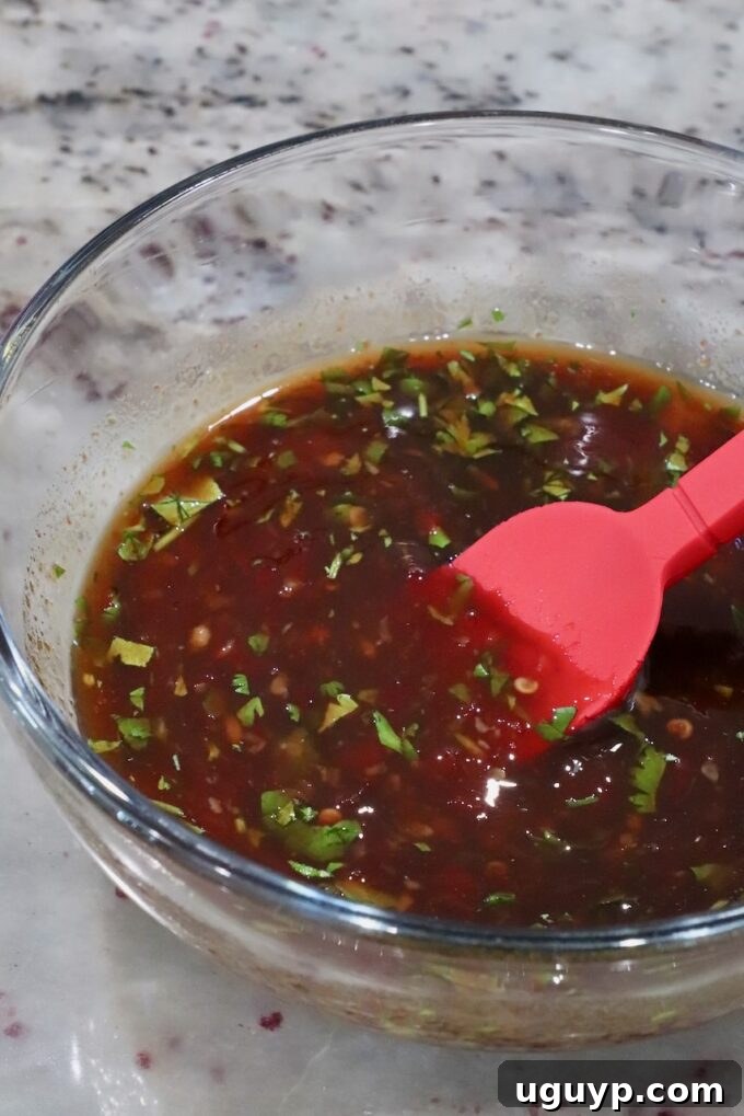 A glistening glass bowl holds a rich, reddish-brown sauce, finely chopped fresh herbs visible within its depth, resting on a sophisticated marble surface. A bright red spatula, poised and ready, is nestled in the bowl.