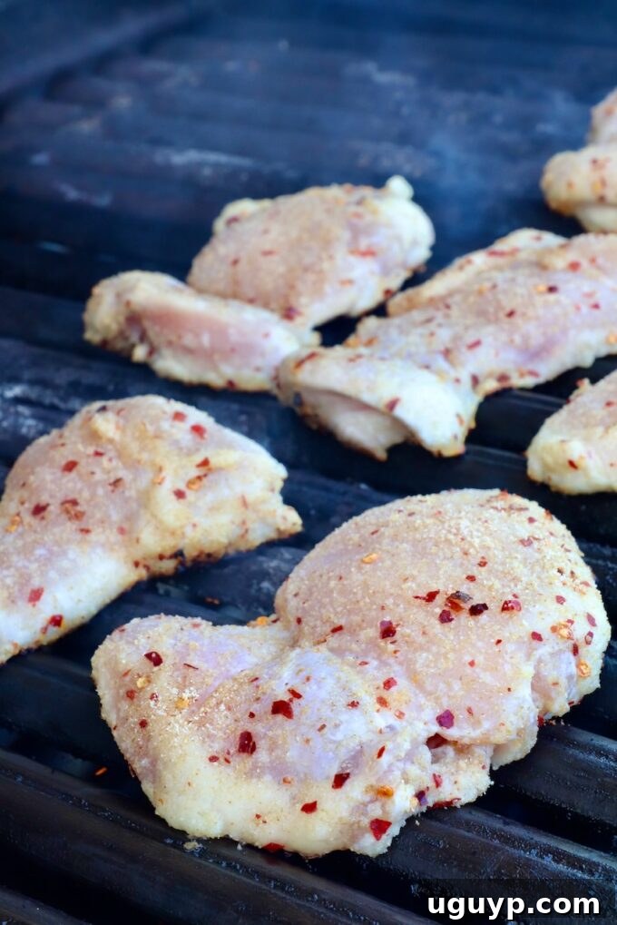 Perfectly seasoned raw chicken thighs, visibly dusted with aromatic spices and a scattering of red pepper flakes, are carefully placed on the hot grates of an outdoor grill, ready to cook.