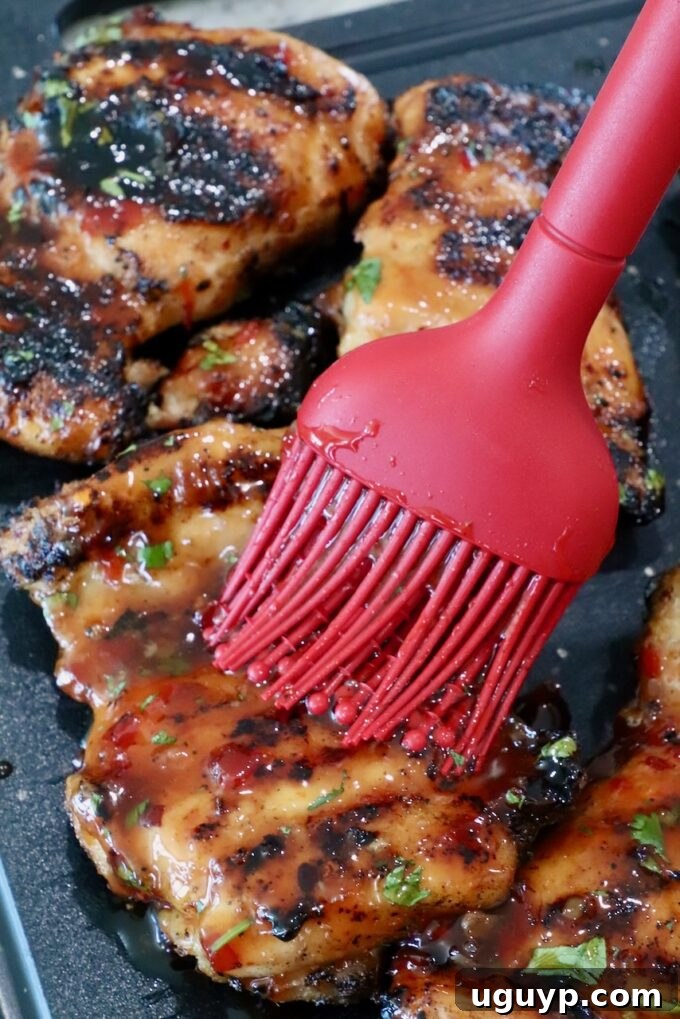Juicy grilled chicken thighs, displaying appetizing charred marks, are meticulously brushed with a thick, glossy sauce using a vibrant red silicone brush, with fresh herbs visible in the background on a dark surface.