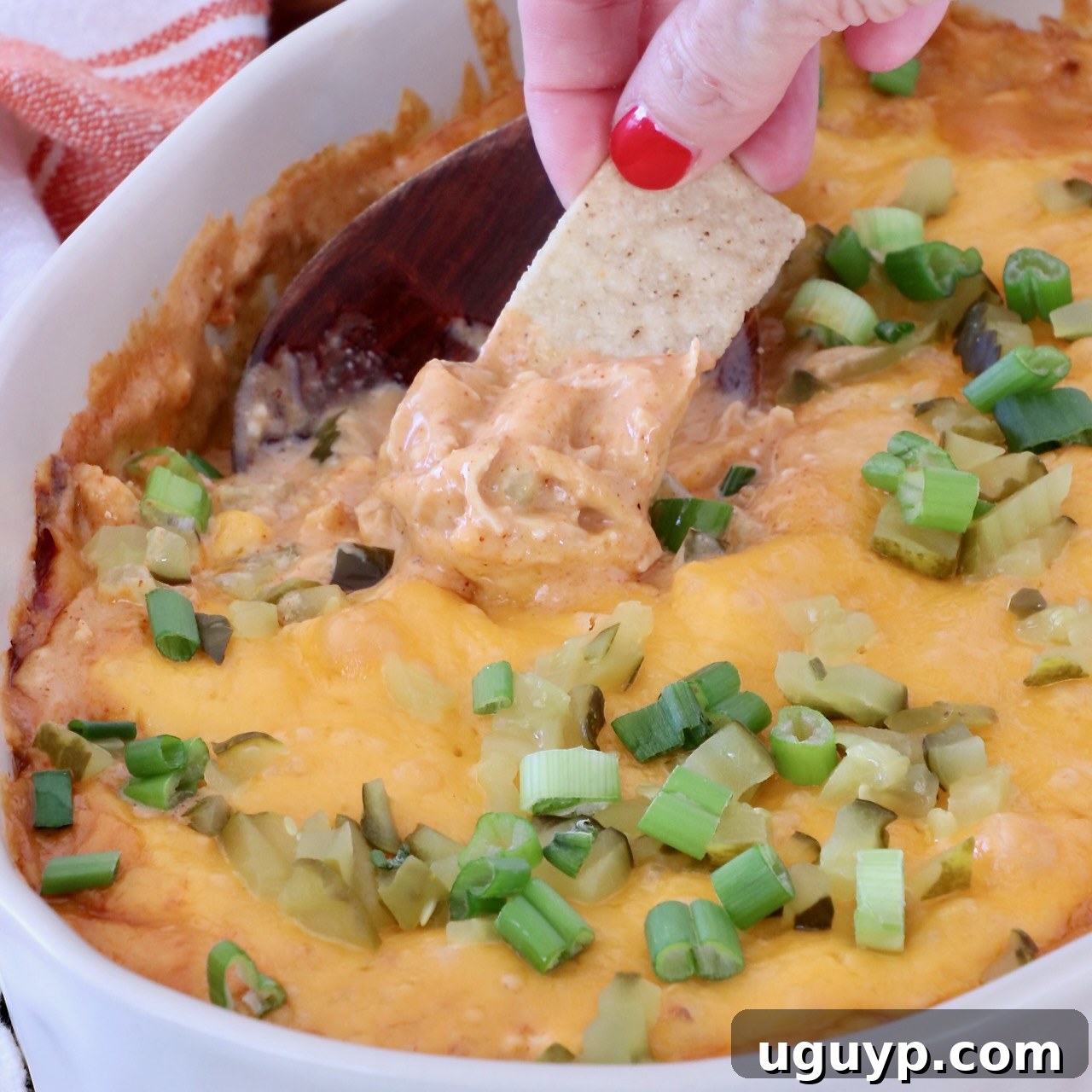 A hand with red nails dips a tortilla chip into a cheesy, baked dip topped with chopped green onions, in a white dish