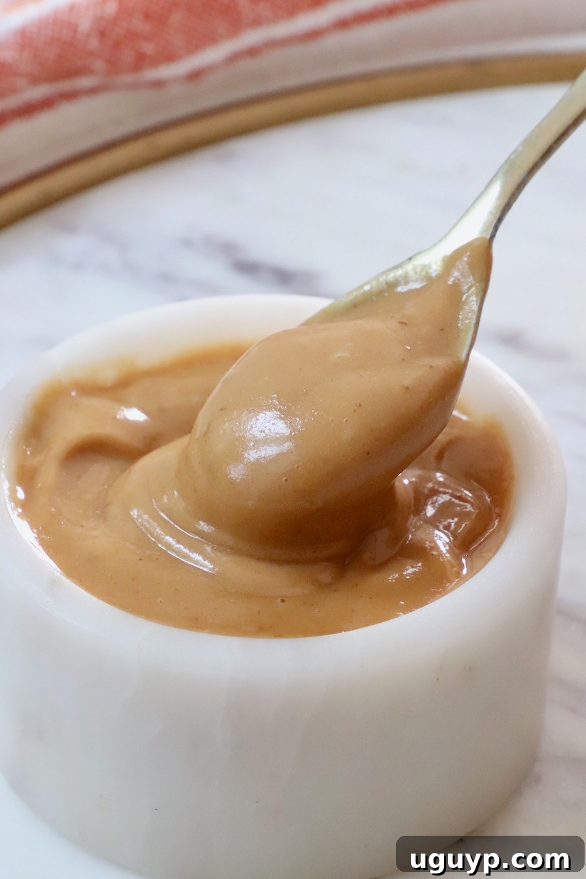 A spoon dips into creamy Thai peanut dressing in a small white bowl on a marble surface, highlighting its rich texture and inviting color.