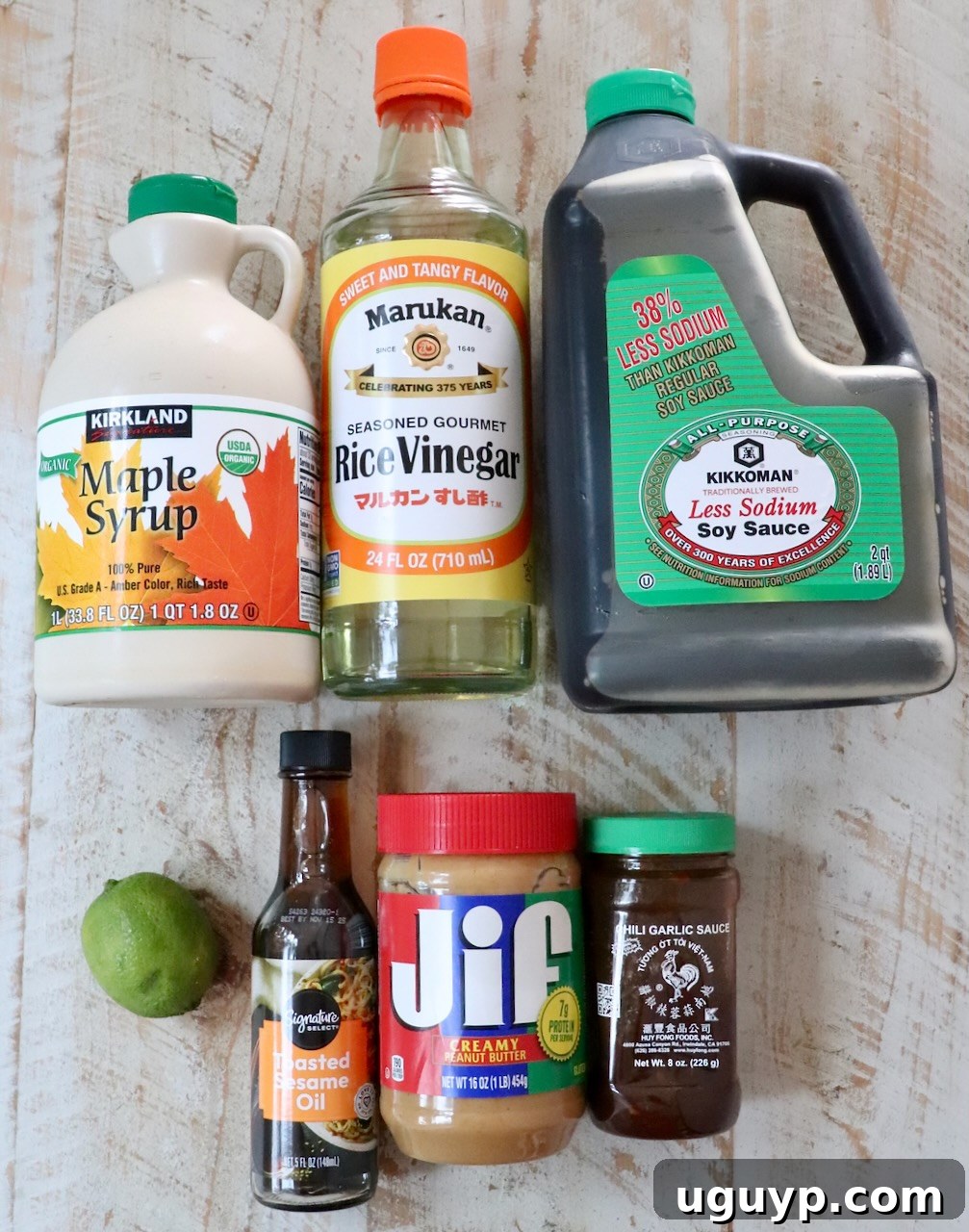 Assorted ingredients for Thai peanut dressing on a rustic wooden table: maple syrup, rice vinegar, soy sauce, sesame oil, creamy peanut butter, chili garlic sauce, and a fresh lime.