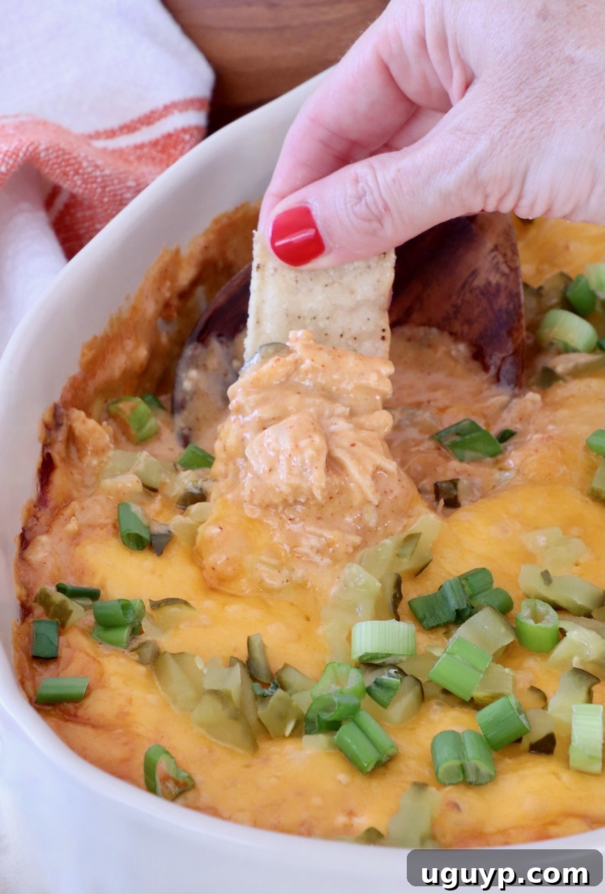 A hand with red nails dips a tortilla chip into a cheesy, baked dip topped with chopped green onions, in a white dish