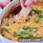 A hand with red nails dips a tortilla chip into a cheesy, baked dip topped with chopped green onions, in a white dish
