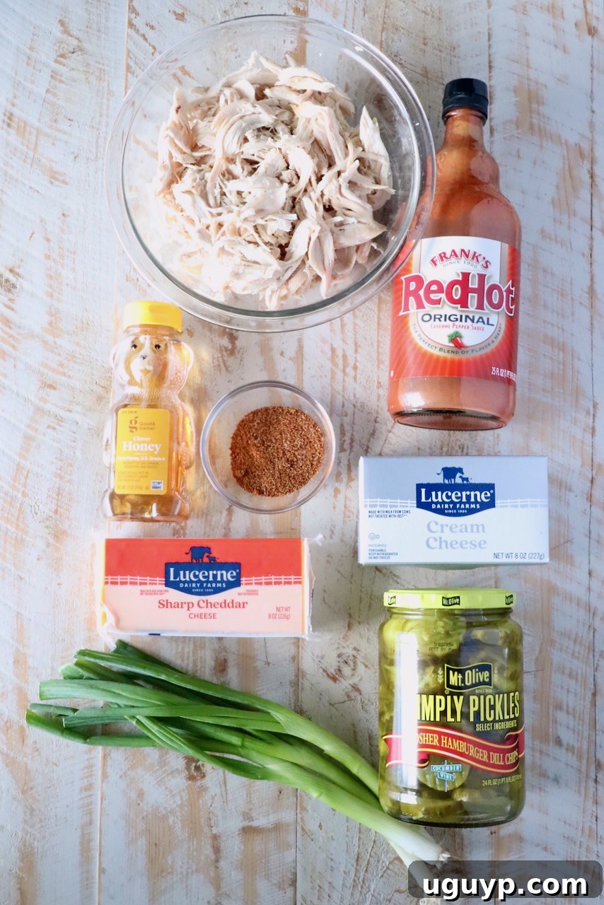 A wooden surface displays ingredients including: shredded chicken, hot sauce, pickles, green onions, honey, cream cheese, cheddar, and seasoning.