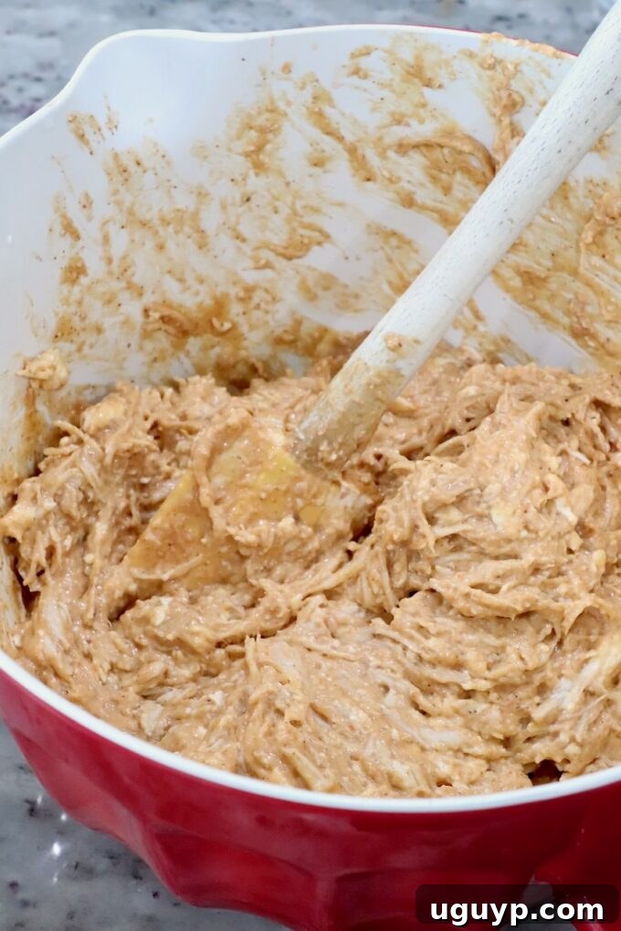 A red bowl filled with a thick, creamy chicken and cheese mixture being mixed with a wooden spoon.