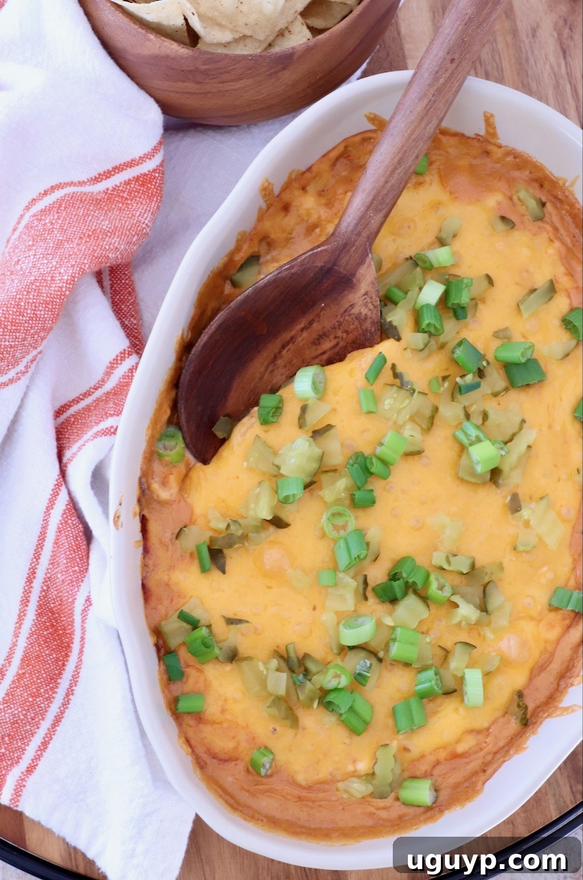 Close-up of a baked cheesy dip in an oval dish topped with green onions and pickles. A wooden spoon rests on top. Tortilla chips in a bowl and a striped towel are nearby.