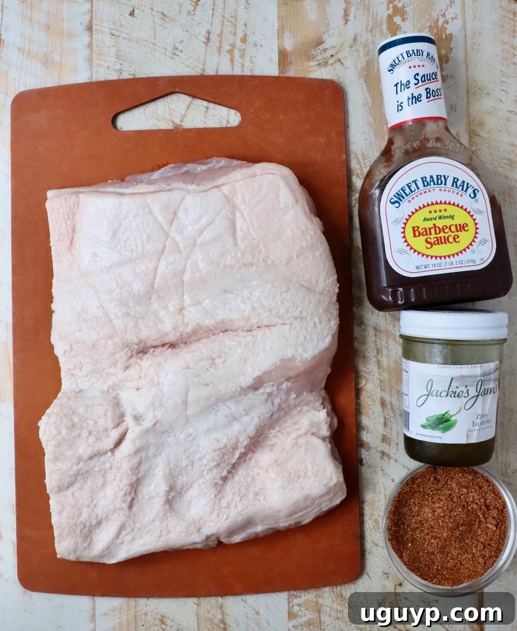 Fresh ingredients for making smoked pork belly burnt ends laid out on a white wooden board, including a block of pork belly, a jar of BBQ seasoning, and bottles of BBQ sauce and pepper jelly.