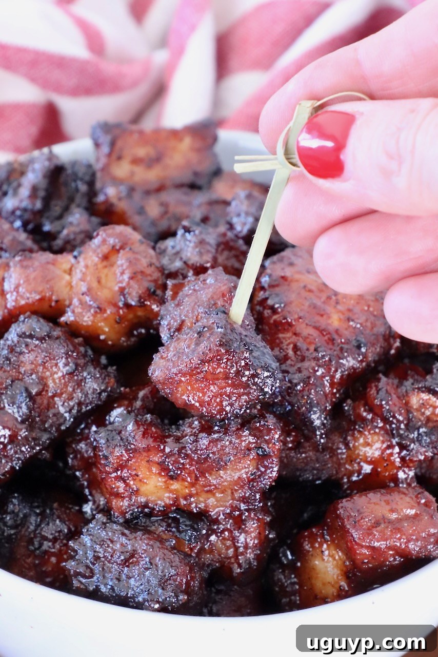A hand with vibrant red nail polish is delicately holding a toothpick, expertly picking up a perfectly glazed and caramelized pork belly bite from a bowl brimming with similar, enticing pieces. A red-striped cloth provides a rustic backdrop, enhancing the visual appeal of the delicious snack.