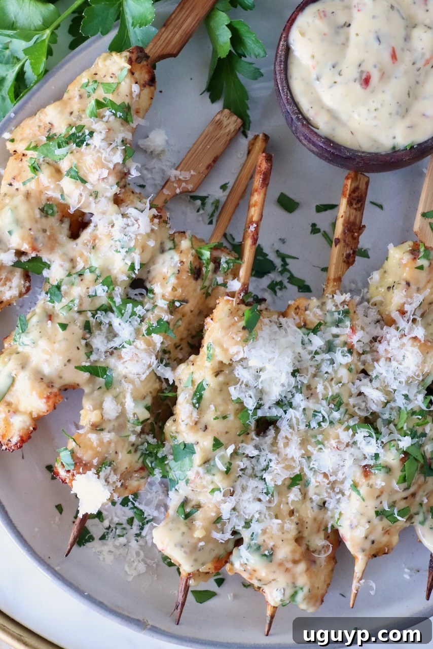 Cooked chicken skewers topped with creamy garlic parmesan sauce and shredded cheese, garnished with chopped fresh parsley. A small bowl of dipping sauce sits alongside.