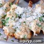 Cooked chicken skewers drizzled with a creamy garlic parmesan sauce, adorned with shredded cheese and fresh herbs, presented on a light-colored plate.