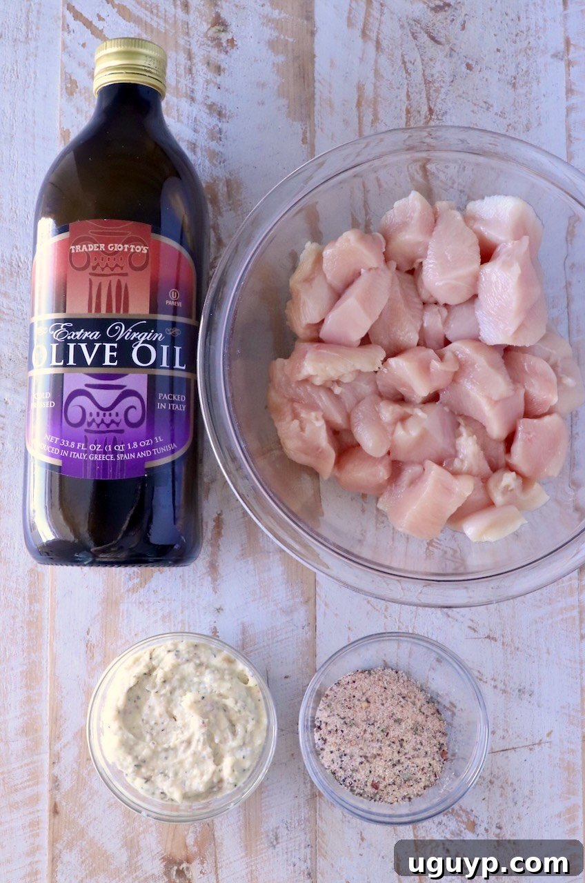 A bottle of olive oil, a bowl of raw diced chicken, and small containers of creamy garlic parmesan sauce and chicken seasoning are arranged on a rustic wooden surface, highlighting the key ingredients for the chicken skewers.
