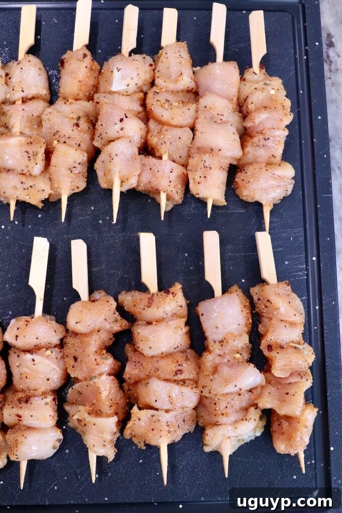 Raw chicken chunks seasoned with spices are neatly arranged on a black cutting board, ready for skewering.