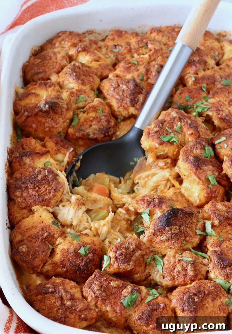 Close-up of a baked buffalo chicken pot pie topped with golden biscuit pieces. Garnished with fresh herbs, a serving spoon rests in the dish. 