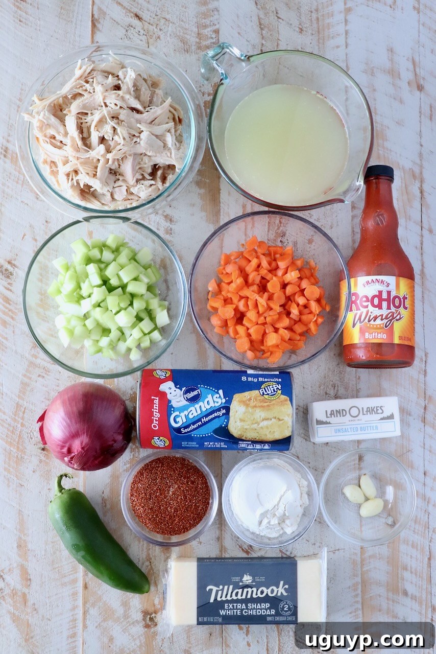 Overhead view of cooking ingredients: shredded chicken, broth, diced celery, carrots, hot sauce, onion, jalapeño, biscuits, butter, seasonings, and cheddar cheese