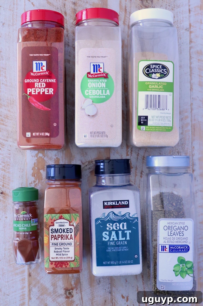 Assorted spice containers, including red pepper, onion powder, garlic, ancho chile, paprika, sea salt, and oregano, arranged on a rustic wooden surface, showcasing the key components of the homemade seasoning.