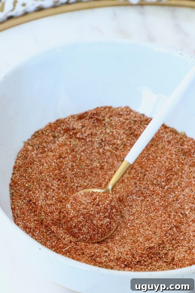 Close-up of a white bowl filled with reddish-brown spice mix. A gold and white spoon rests on top, inviting you to mix and use this flavorful homemade chorizo seasoning.