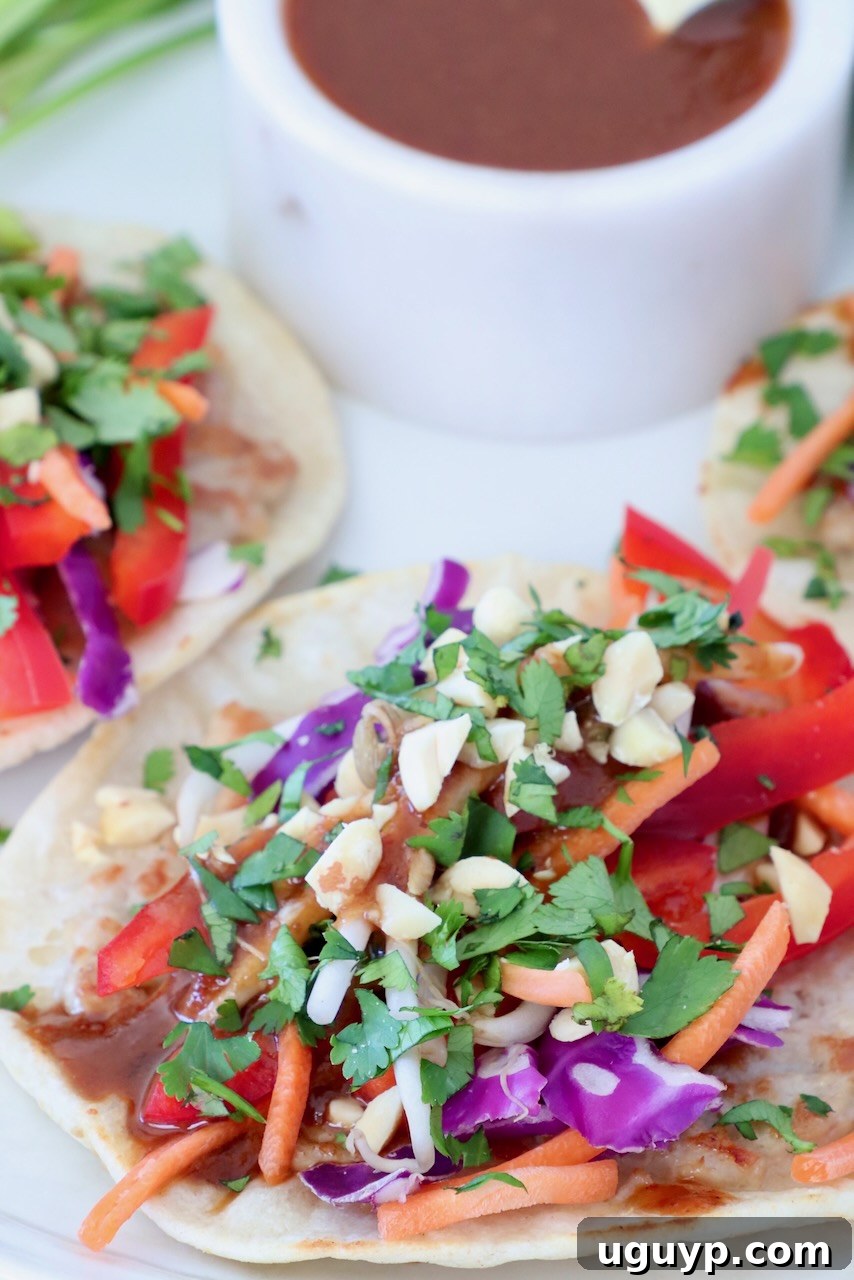 Close-up of pad thai tacos with vibrant toppings like red peppers, carrots, purple cabbage, and fresh cilantro, next to a bowl of sauce, highlighting their colorful appeal and deliciousness.