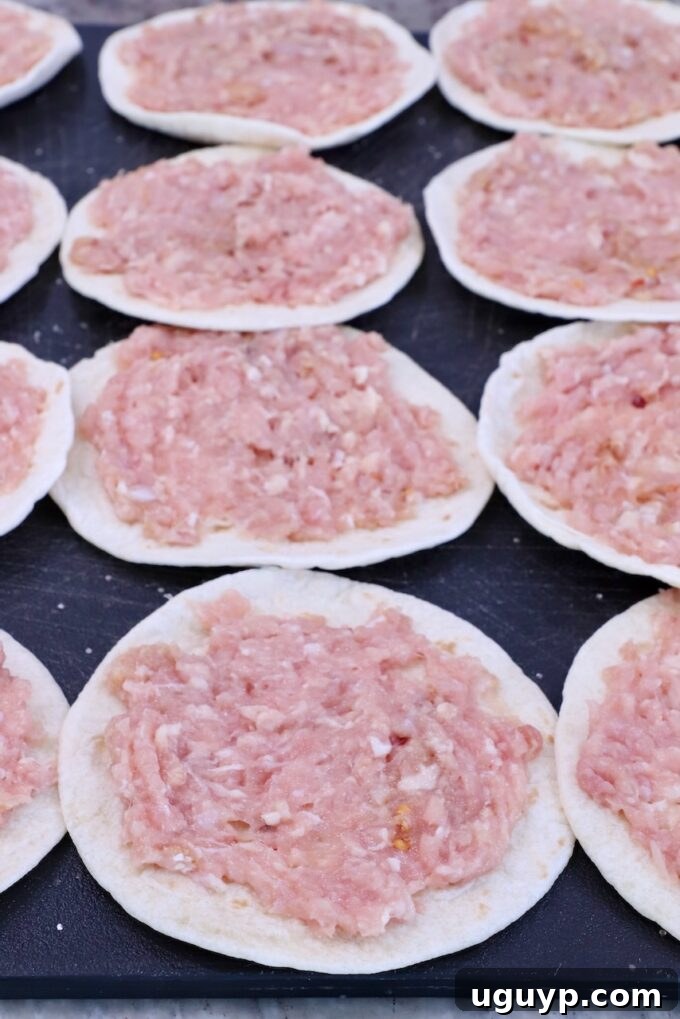 Raw ground chicken is spread evenly on small, round tortillas laid out on a dark surface, prepped for smashing and cooking.