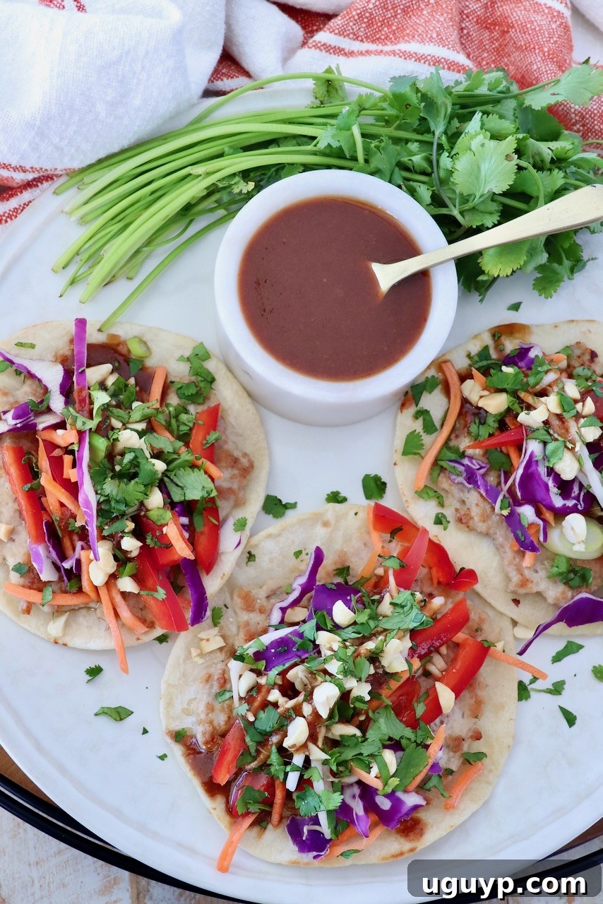 Plate of colorful pad thai tacos topped with vibrant red peppers, purple cabbage, carrots, and fresh cilantro. A ramekin of sauce and cilantro garnish beside, inviting you to dig in.