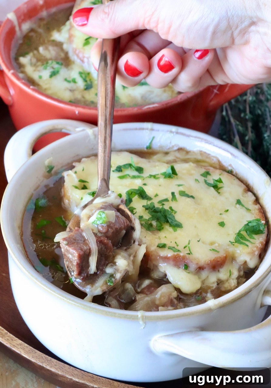 A steaming bowl of French onion beef stew, adorned with golden-brown melted Gruyere cheese and fresh herbs. A hand delicately lifts a spoon, showcasing a tender piece of beef and a caramelized onion.