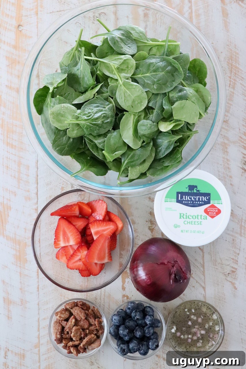 Assortment of fresh ingredients for spinach strawberry salad laid out on a white wooden board, including baby spinach, sliced strawberries, blueberries, red onion, pecans, and ricotta cheese.