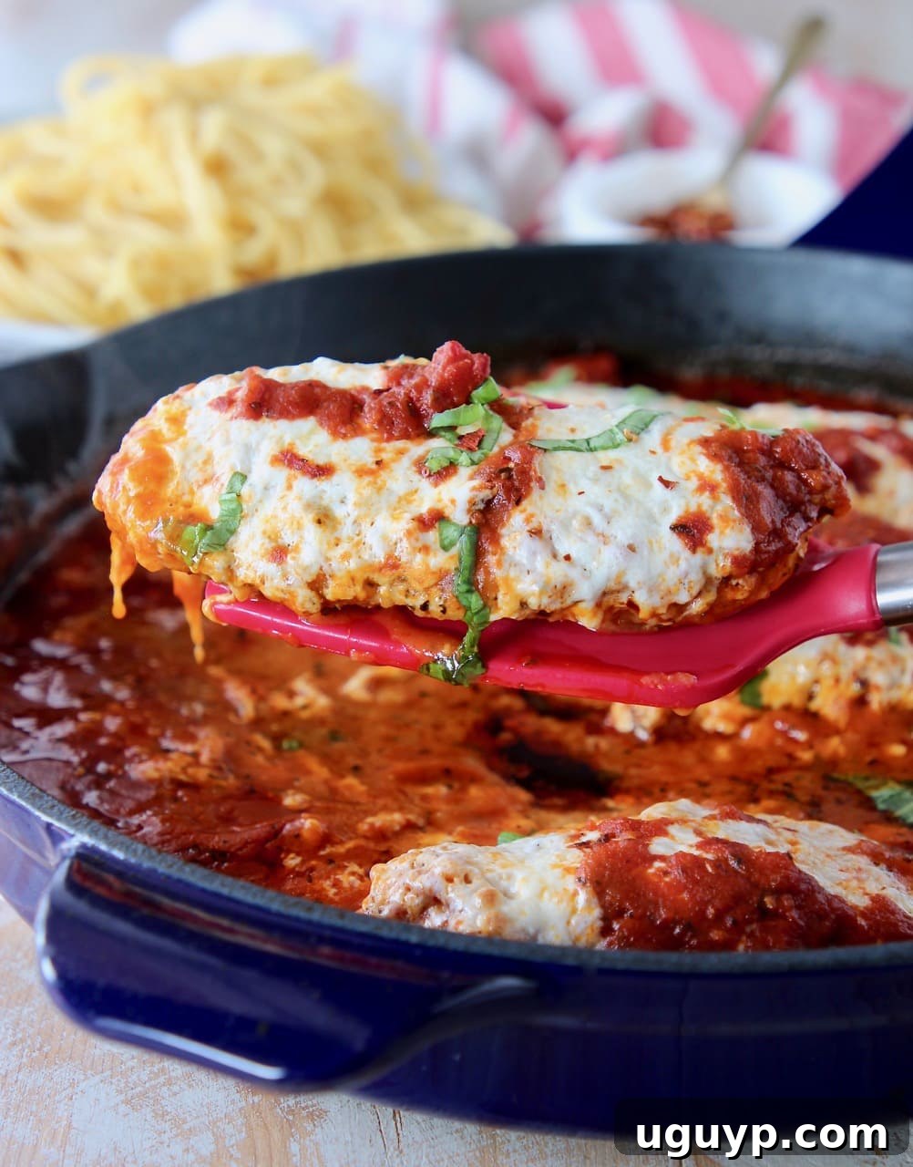 A vibrant red spatula lifting a perfectly baked piece of gluten-free chicken parmesan from a large blue skillet, showcasing its golden crust and melted cheese.