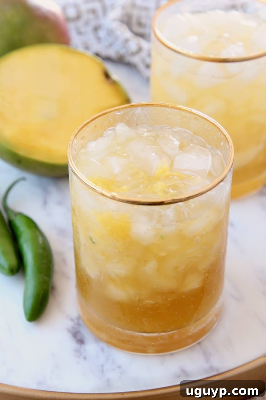 Two spicy mango margaritas in glasses, garnished with chili and lime, next to fresh sliced mango on a marble tray.