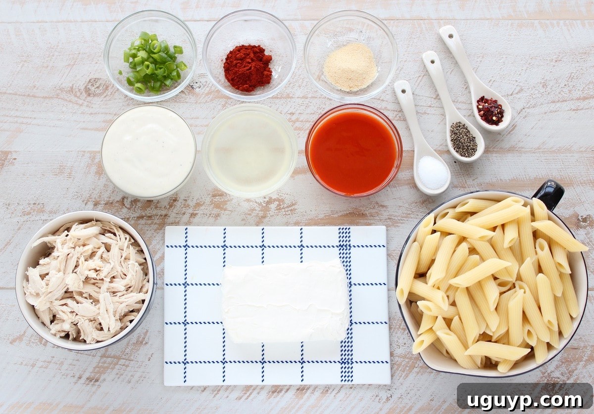 Fresh ingredients for Buffalo Chicken Pasta arranged on a white wooden board, including shredded chicken, cream cheese, buffalo sauce, chicken broth, spices, and pasta.