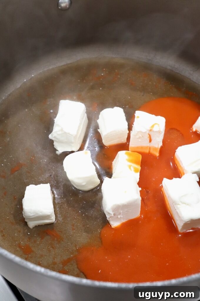 Cubed cream cheese melting in a large pot with buffalo sauce and chicken broth, forming the base of the creamy sauce.